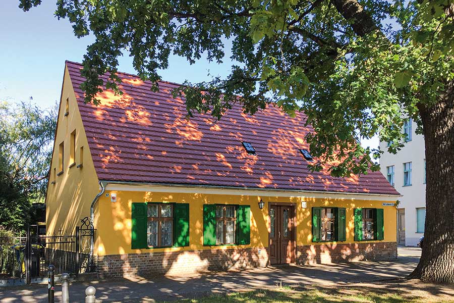 Ein einstöckiges gelbes Haus mit hohem Dach und grünen Fensterläden, davor ein großer Baum