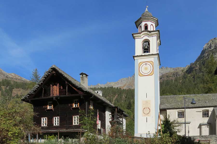 Ein dunkles altes Bauernhaus aus Holz steht neben dem hellen Kirchturm, dahinter Berge